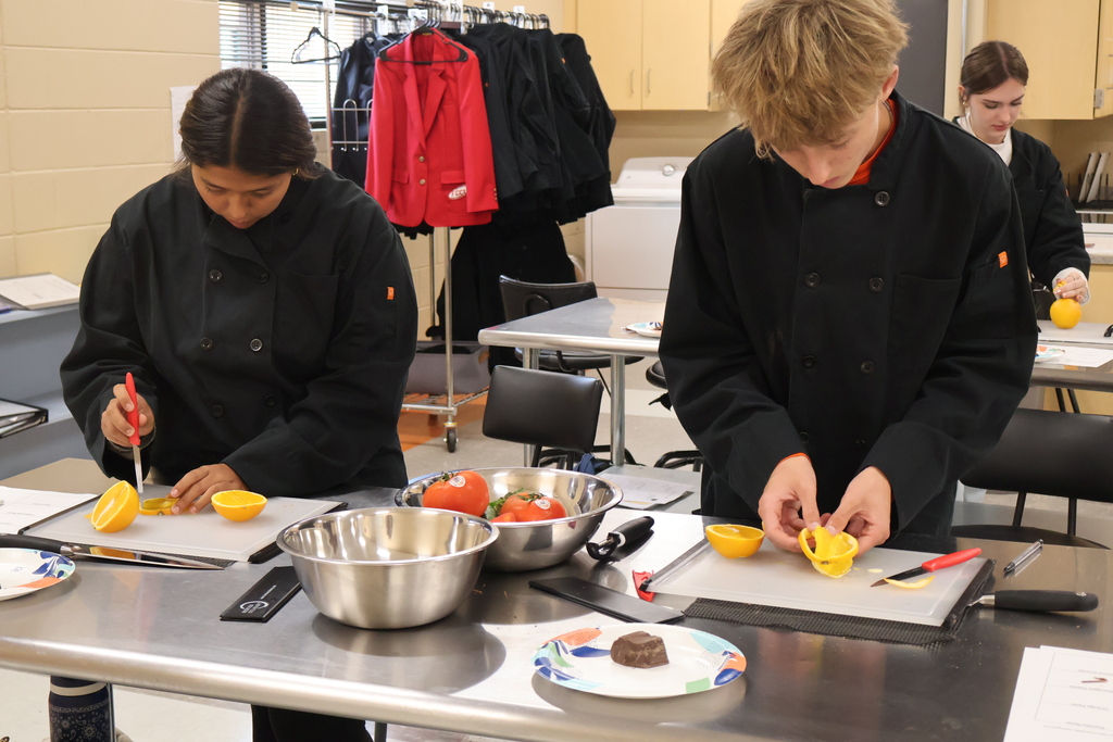 There are several students all standing at their silver tables with bowls of fruits and vegetables as they are learning to cut orange peels to use as a garnish. 