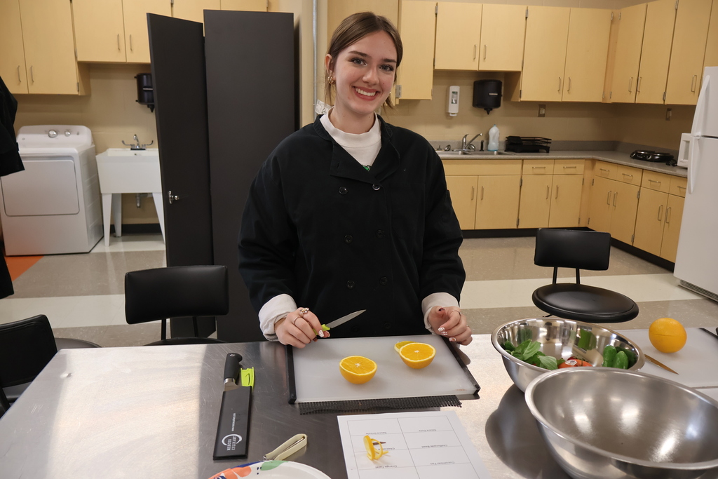 A student is standing at a silver table as she is practicing cutting an orange peel to use as a garnish. 