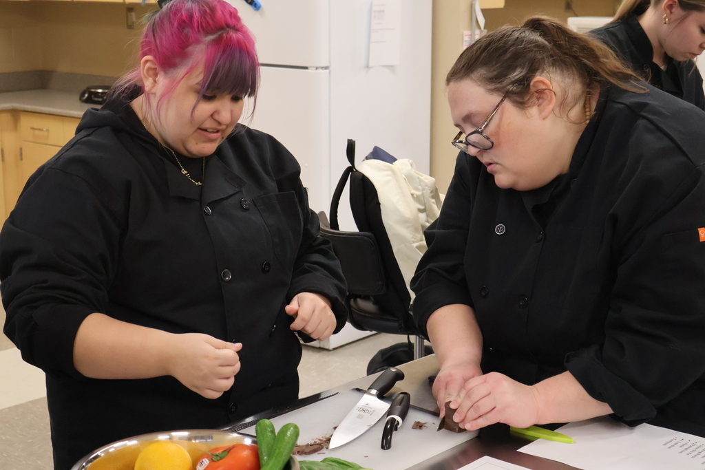 The teacher is helping a student at her table learn how to properly cut chocolate into curls. 