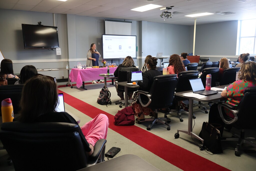 There are several people seated at tables with their laptops in front of them with one person standing up at the front of the room speaking to them with a board beside her.