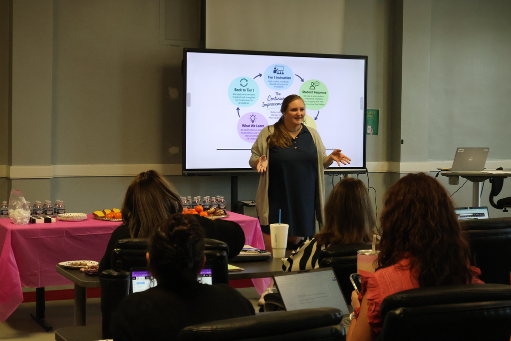 There are several people seated at tables with their laptops in front of them with one person standing up at the front of the room speaking to them with a board beside her.