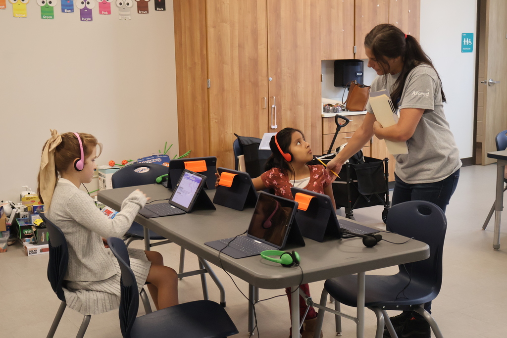 Two students are sitting at a table while wearing headphones and working on a computer and a woman is standing behind one of them. 