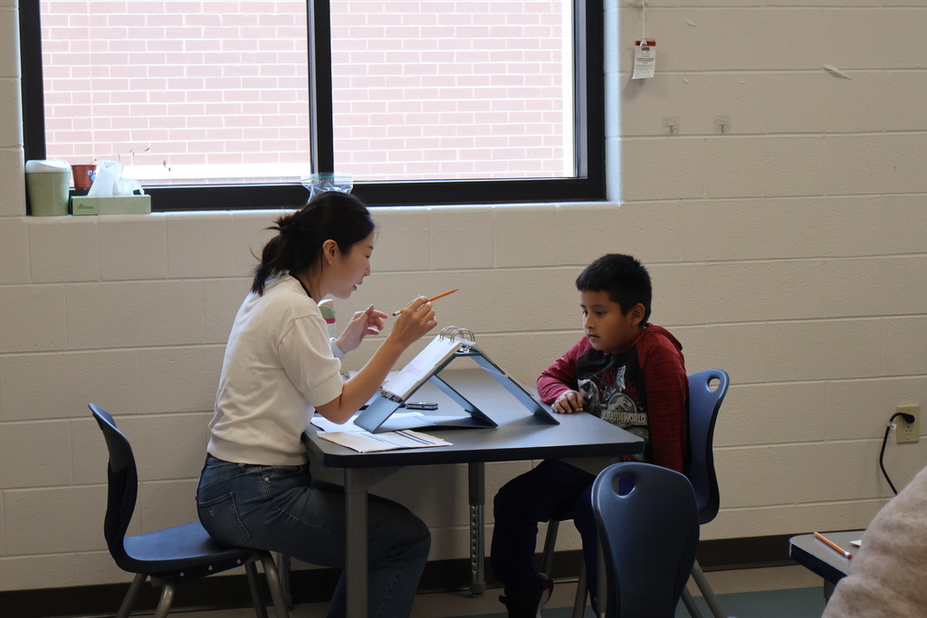 A woman is sitting at a table with a student while they work on reading together. 