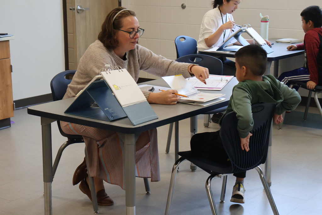 A woman is sitting at a table with a student and she is reaching across the table to give him a fist bump. 