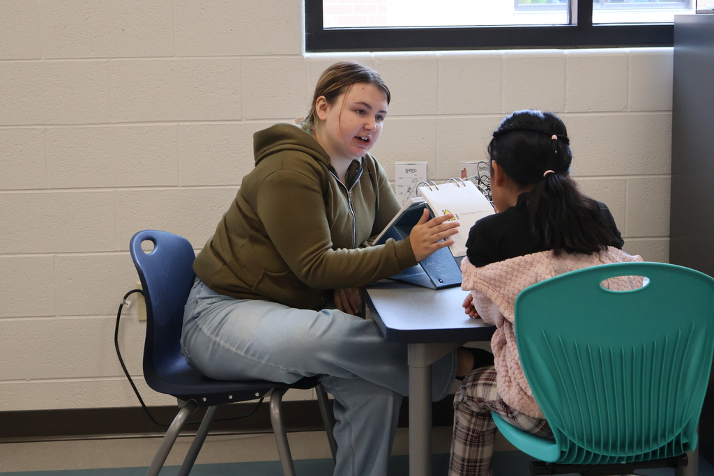 A woman is sitting at a table with a student while they work on reading together. 