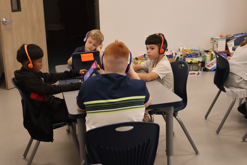 Several students are sitting at a table wearing headphones while also working on a computer. 