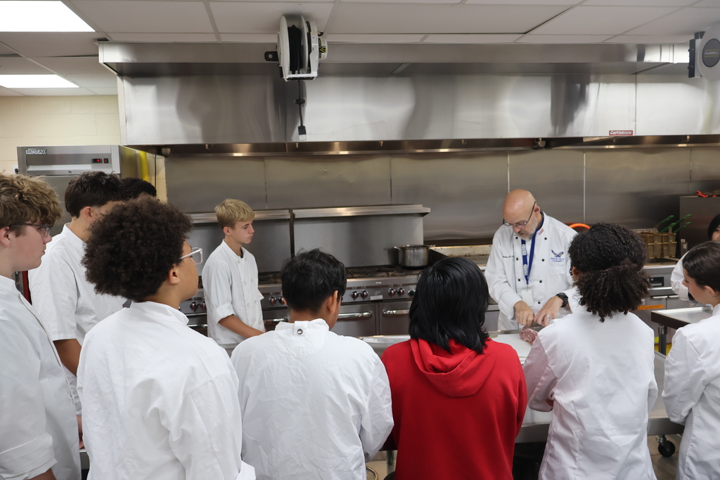Students are gathered around a metal table while the teacher talks to them about pork. 