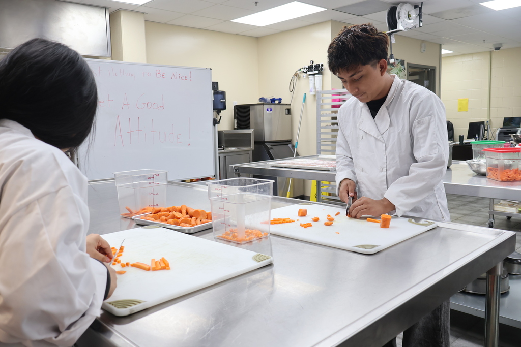 Two students are standing at a table in the Culinary Arts class cutting up carrots. 