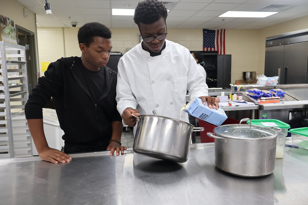 Two students are adding cream to a large pot of potatoes. 