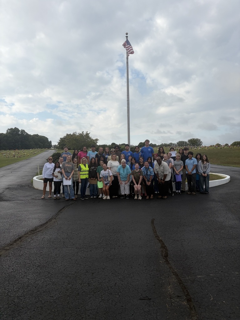 This is a large group picture of students and teachers and they are standing in front of the flag pole at the cemetery. 