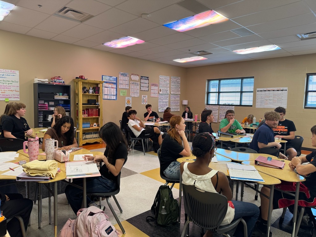 Honors English II students seated at grouped desks in classroom discussing Frankenstein and The Metamorphosis, working on papers and engaging in literary analysis conversation.