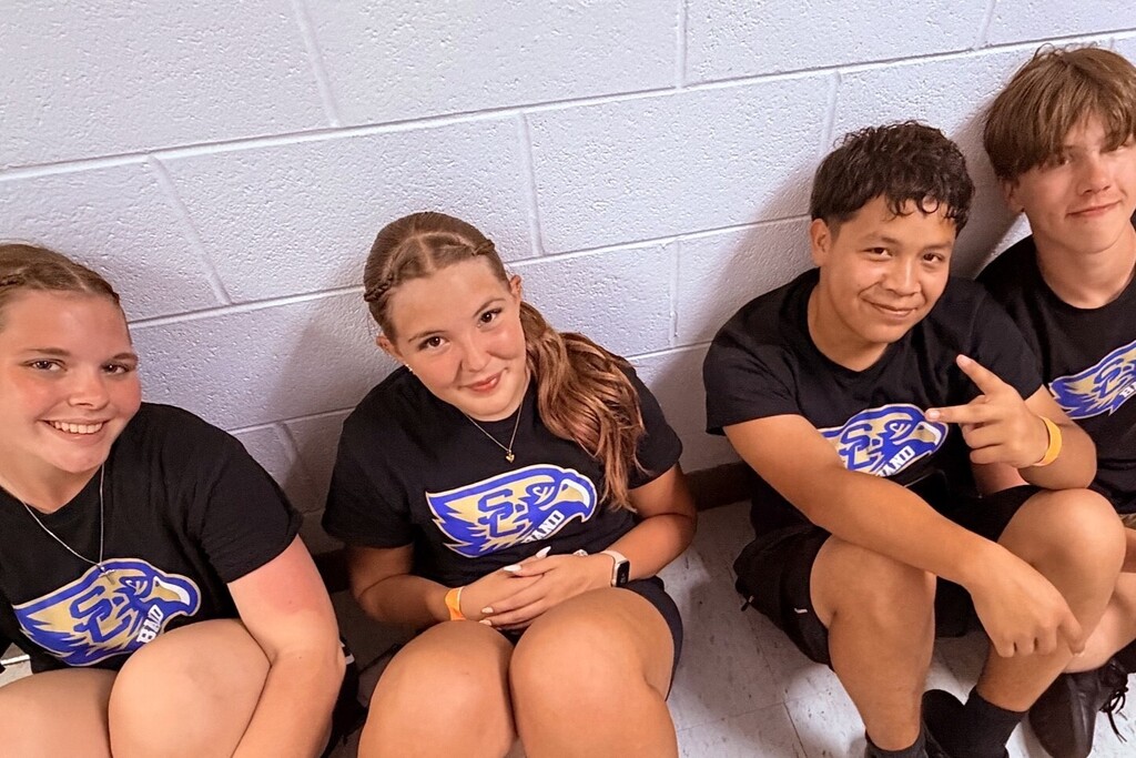 Four band students are sitting in the hallway on the floor against the wall. 