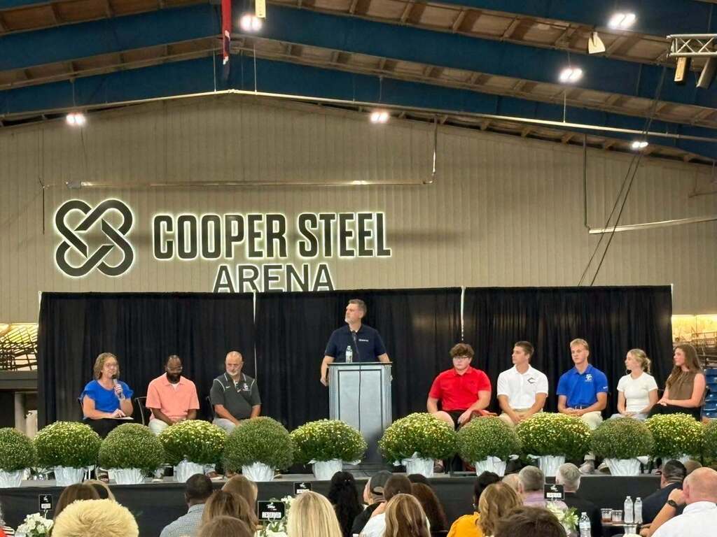 A man is standing at a podium on the stage inside Cooper Steel Arena and is speaking to the crowd with others who are also seated on stage. 