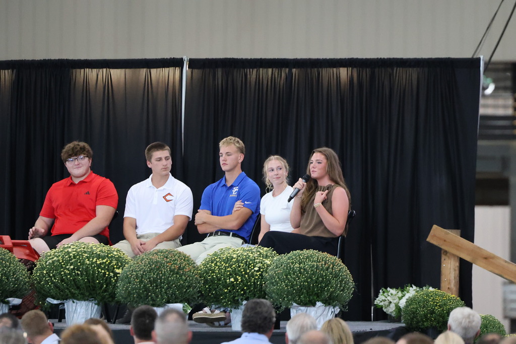 A student sitting on stage is speaking to the crowd. 