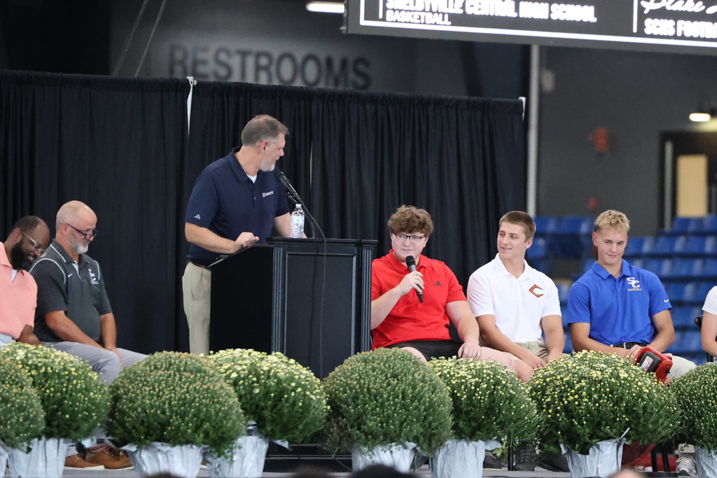 A student sitting on stage is speaking to the crowd. 