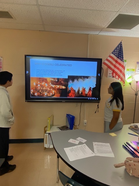 Two students are standing in the classroom beside a touchscreen tv ready to give their presentation to the class.