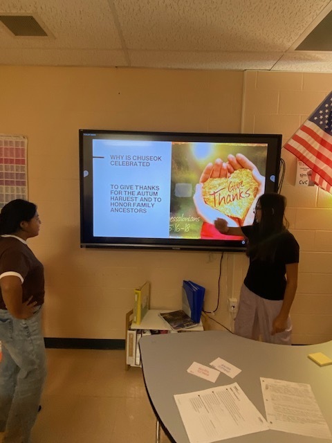 Two students are standing in the classroom beside a touchscreen tv ready to give their presentation to the class.