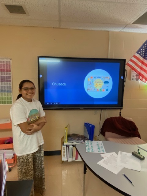 A student is standing in the classroom beside a touchscreen tv ready to give her presentation to the class. 