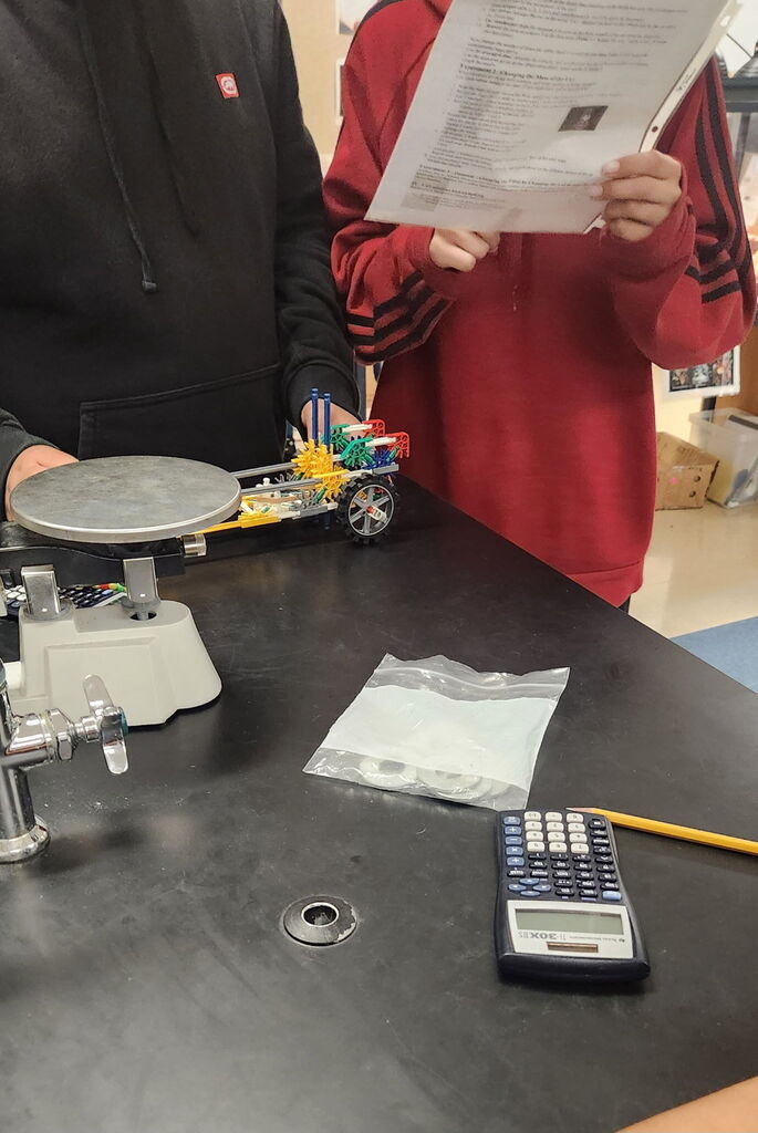Students are standing together by a table and looking at a piece of paper with their K'nex car in front of them. 