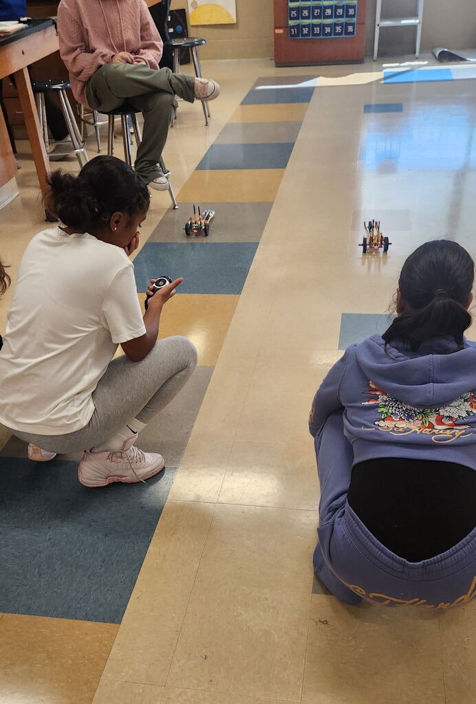 Two students are watching their K'nex cars roll across the classroom. 