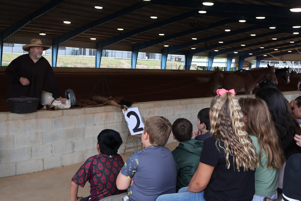 A man is explaining homesteading to a group of students who are sitting on bleachers in front of him. 