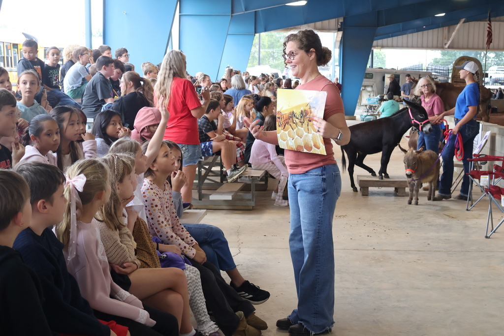 A woman is holding a picture of a live honeybee on a honeycomb while talking to students who are seated on bleachers in front of her. 