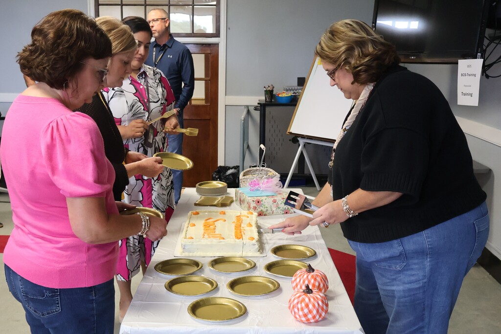 A woman is cutting the cake while three other women are standing in front of it holding plates to hand out. 