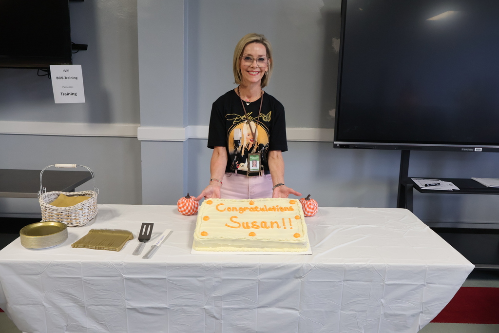 A woman is standing behind the decorated table and is holding up the white and orange cake. 