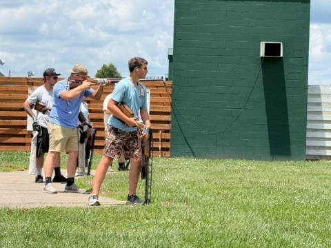Group of four trap shooters on grassy range beside tall dark-green wall, with three aiming firearms and one holding firearm pointed downward.
