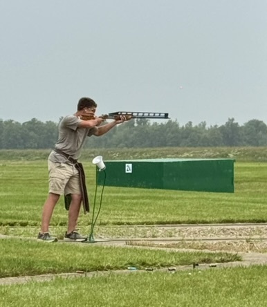  Tanner aiming shotgun at outdoor trap shooting range beside green barrier marked "2A" with grassy fields and trees in background.