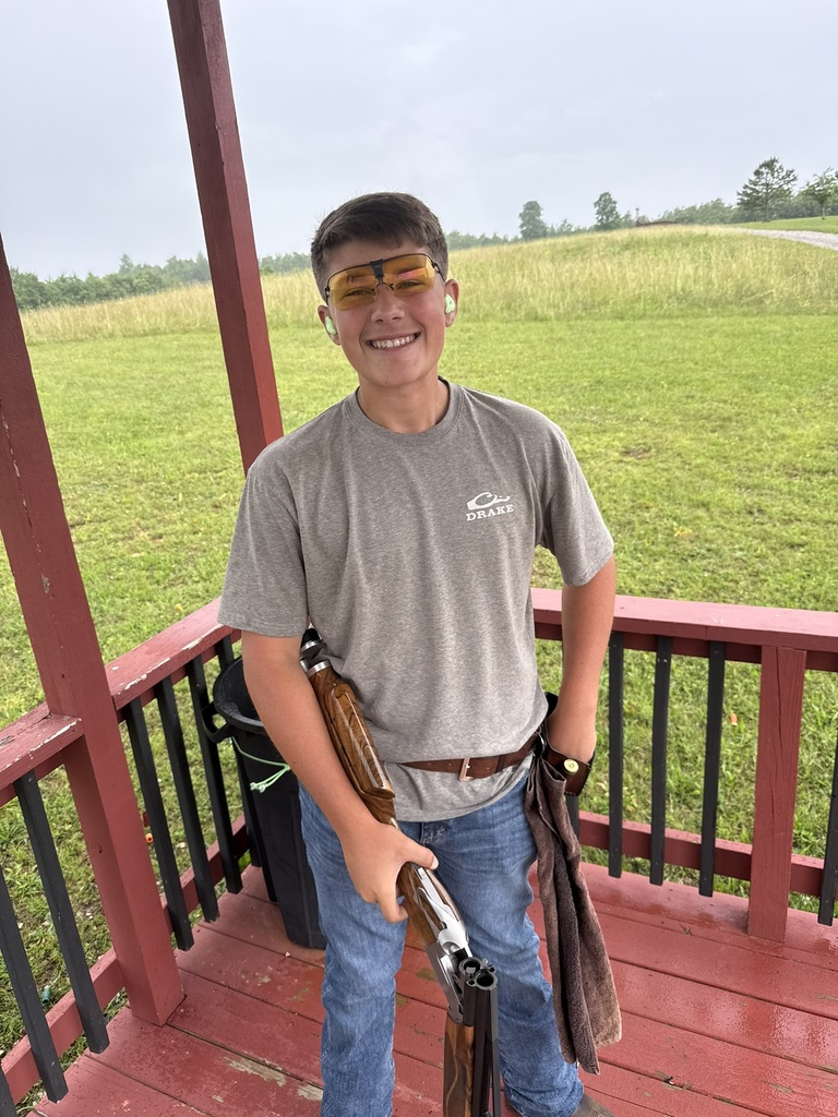 Cascade High School student Tanner standing on red shooting platform holding shotgun, wearing yellow-tinted protective glasses, green earplugs, and gray Drake t-shirt at trap shooting range.