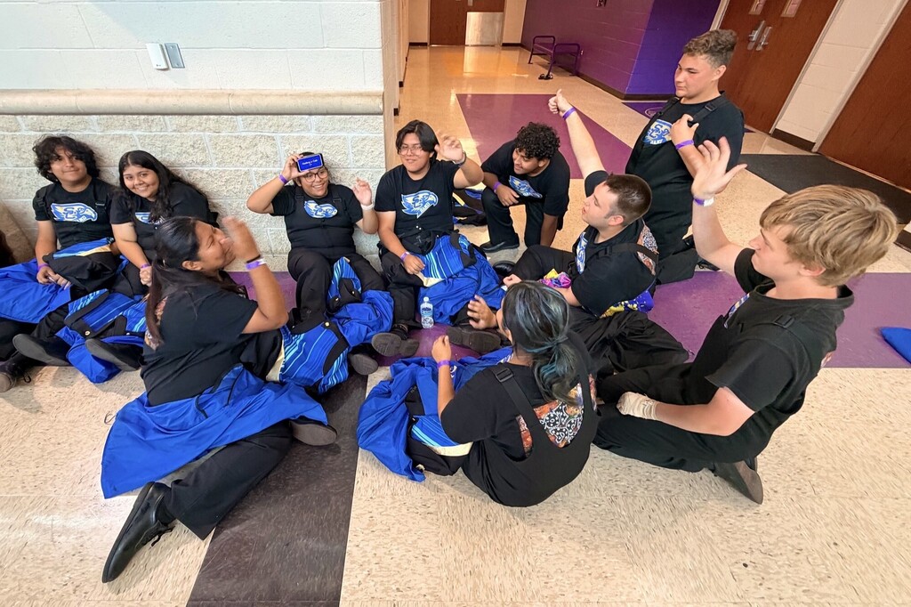 Several members from the marching band are sitting on the floor in the hallway playing a game together while waiting on their performance. 