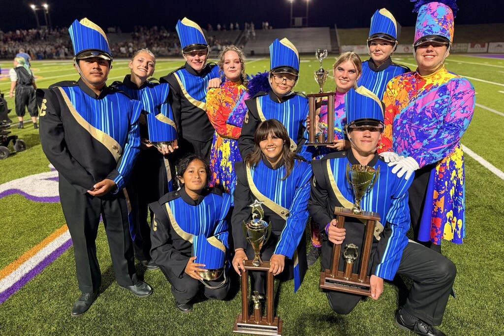 There are several students from the marching band all posing for a group picture on the football field while some are holding trophies. 