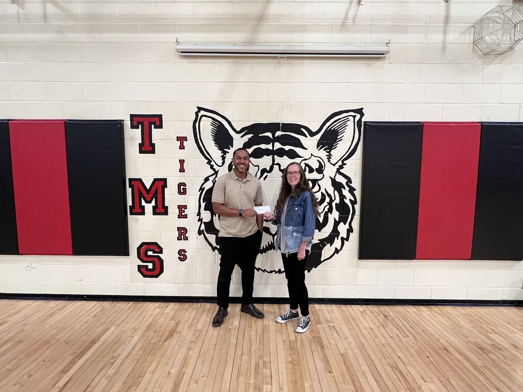 A man and a woman are posed for a picture in the gym in front of the school's mascot that is painted on the wall. 