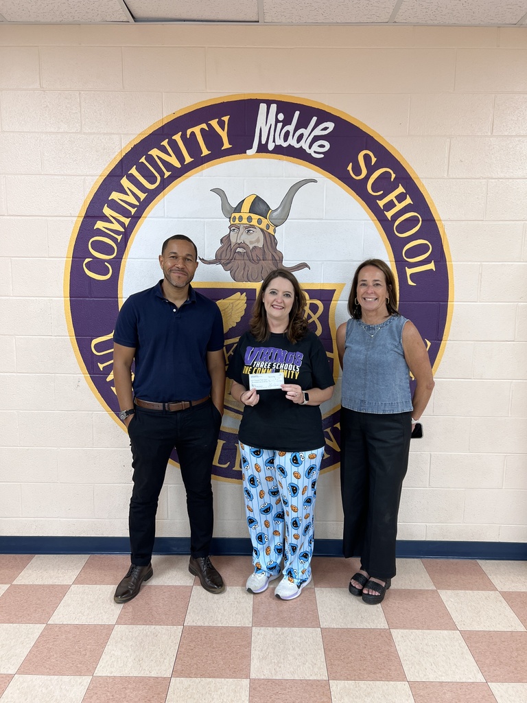 A man and two women are posed for a picture in the hallway in front of the school's mural of their logo and one woman is holding up a check. 