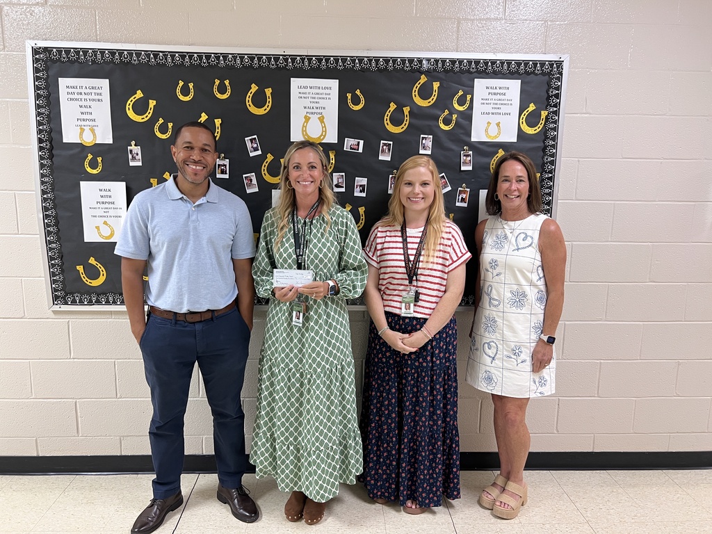 Four people are posed for a group picture in the hallway of the school while one is holding a check. 