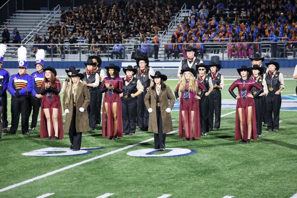 Members of the marching band are standing on the football field during their performance wearing cowboy and cowgirl related apparel. 
