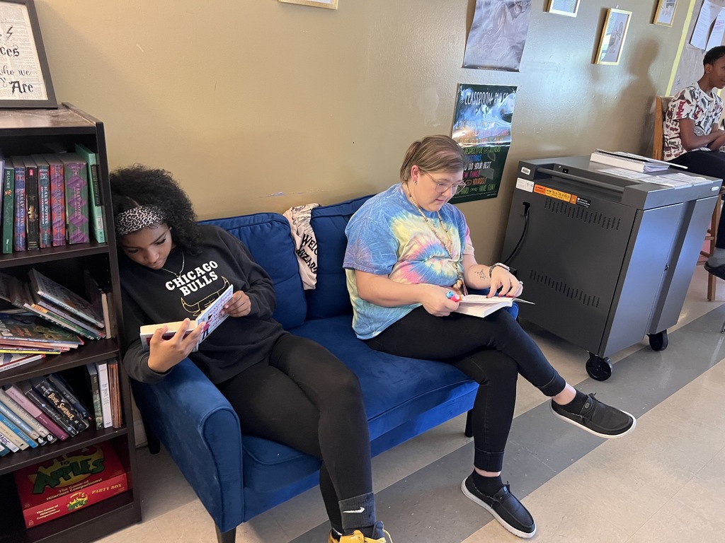 Two students are sitting on a blue couch reading books. 