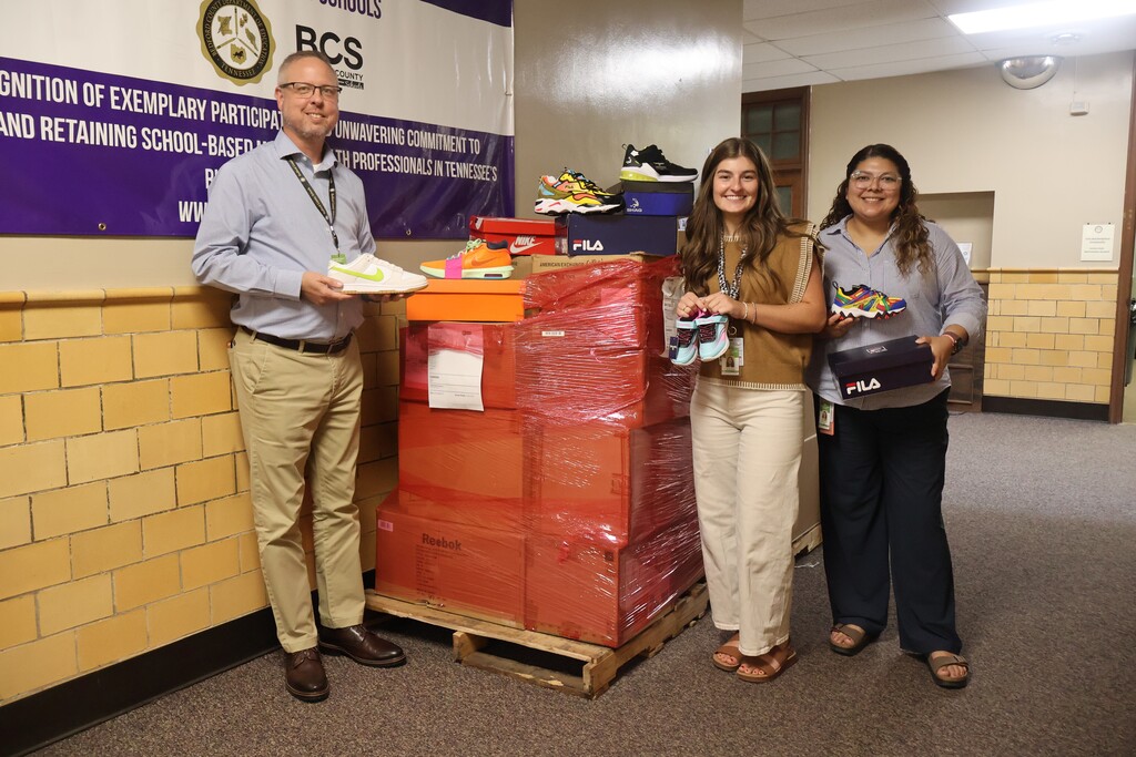 A man and two women, all holding tennis shoes, are standing beside a pallet of donated shoes. 