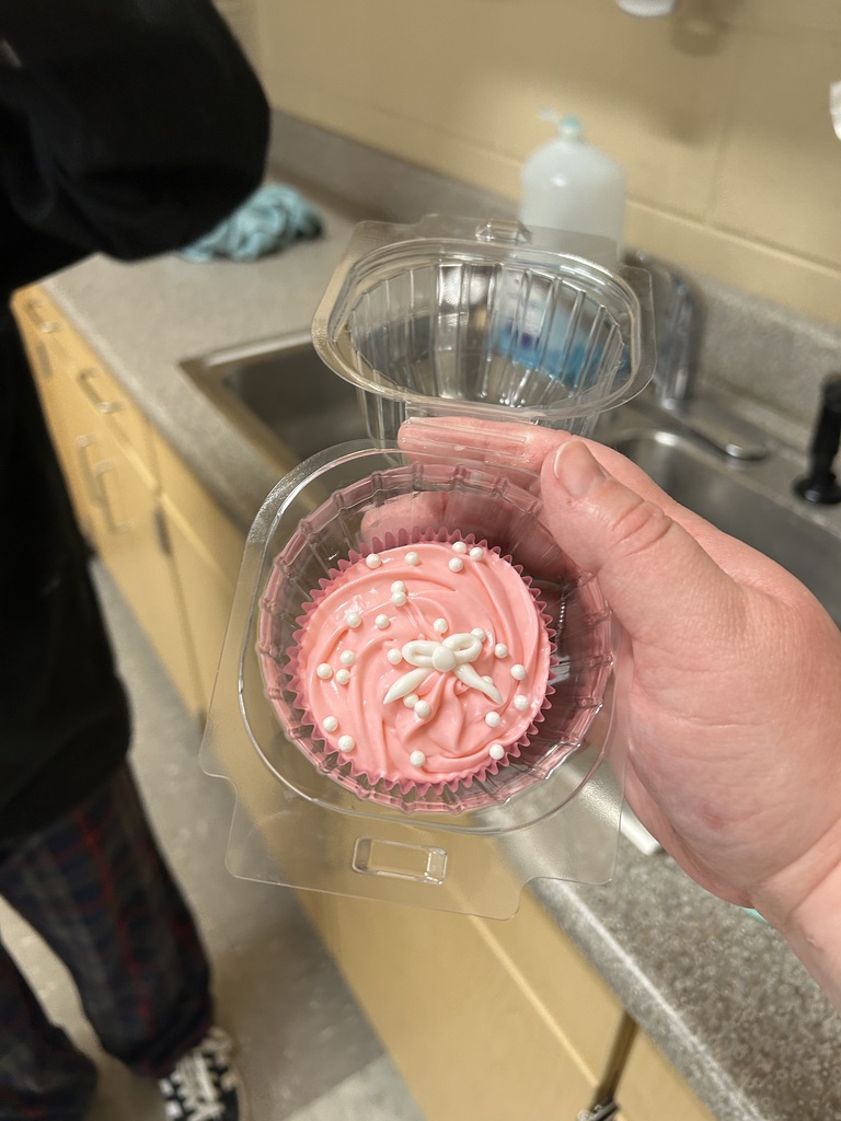 Hand holding open clear container with red velvet cupcake topped with pink buttercream frosting, white pearls, and handmade white candy bow in school kitchen.