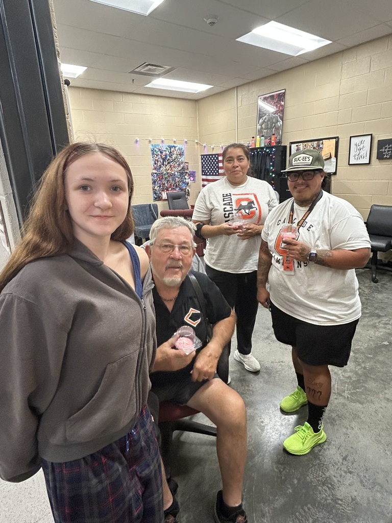 Four Cascade High School custodians posing together indoors, one seated holding pink cupcake container while three stand nearby, celebrating National Custodian Day.