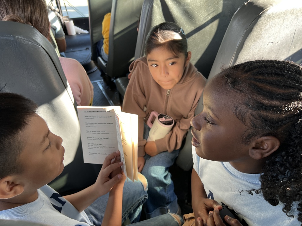 One student is sitting in his bus seat while two other girls are listening to him read from their seats. 