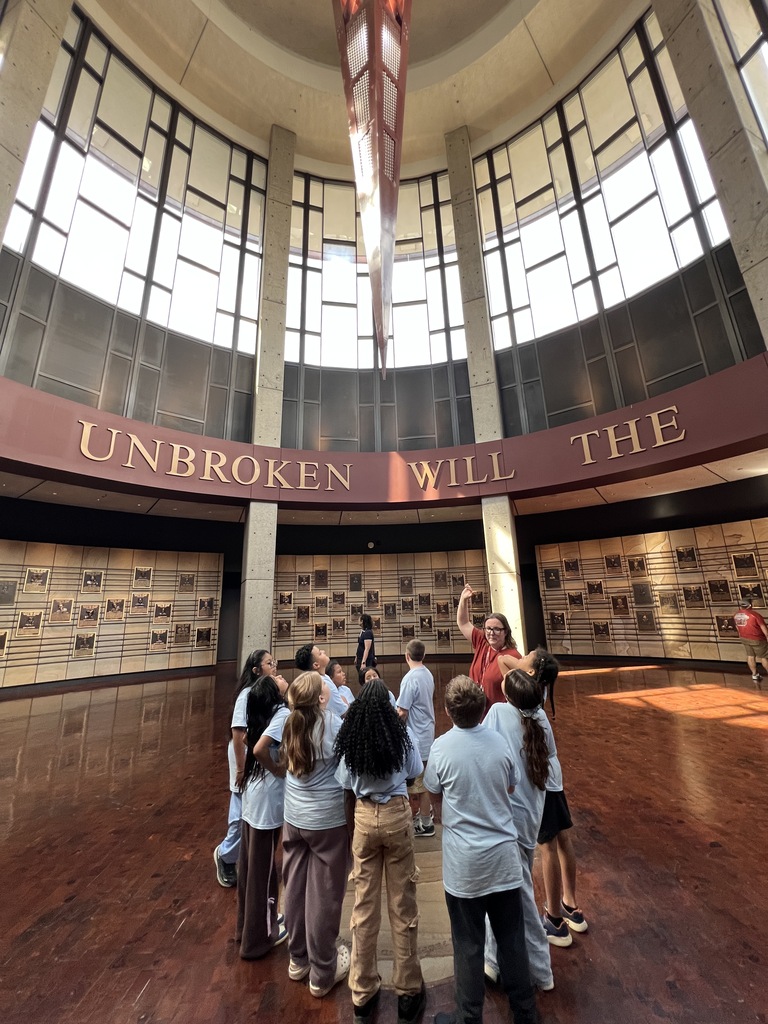 There is a group of students and some are looking up at the tall ceiling while a woman is talking to them. 