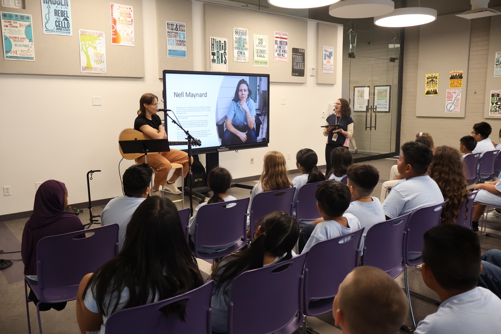 There is a large group of students all sitting in purple chairs and they are listening to two women who are speaking. One of them is sitting on a stool holding a guitar. 