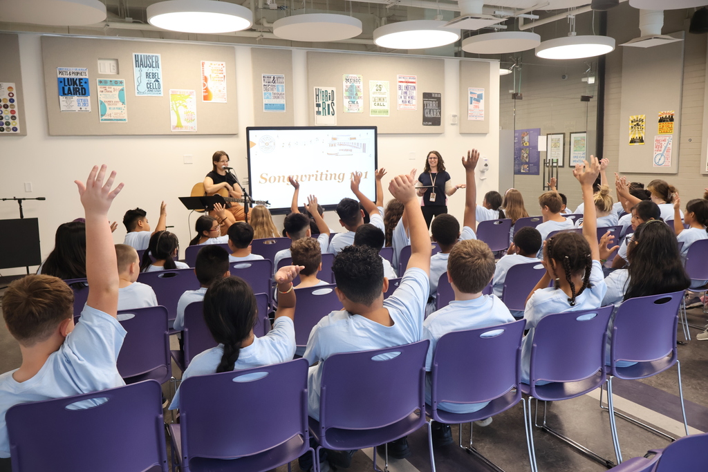 There is a large group of students all sitting in purple chairs and many have their hands raised as two women are standing up front asking questions. 