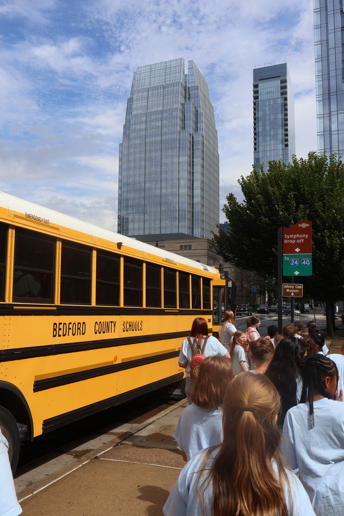 Students are in a line waiting to get on their bus with some tall buildings in the background. 