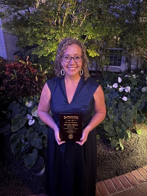 Principal Meredith Gilliland is holding a plaque outside by some flowers.