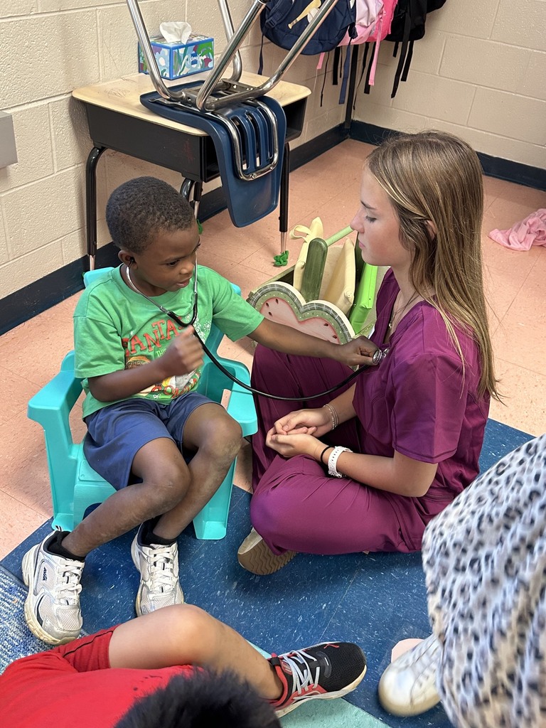 A student is sitting in a chair beside a HOSA student and is using stethoscope to listen to her heartbeat.