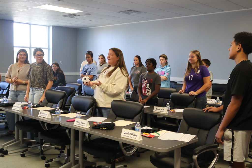 A student is holding a ball and talking while other students around her are listening and watching. 