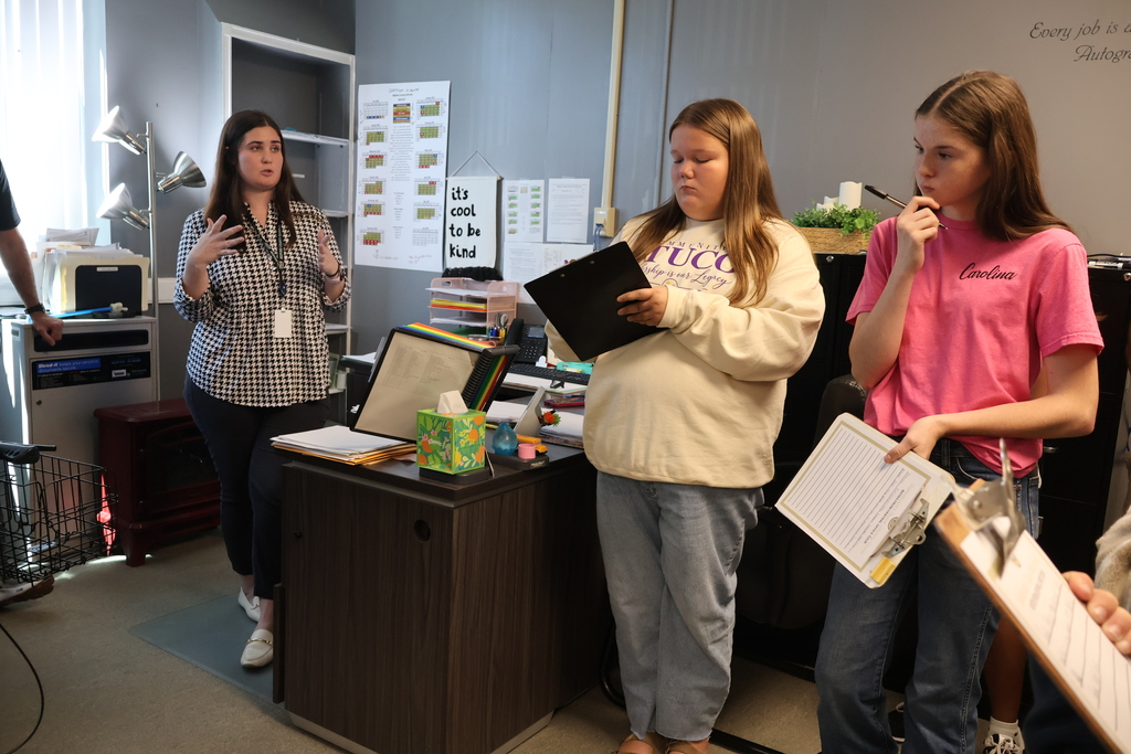 Several students are standing and taking notes on a clipboard while an adult is talking to them all. 